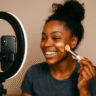 GRWM or GRWMeh? Why the TikTok Beauty Trend Is Both Comforting and Cringe 5 A smiling Black American woman applies foundation with a makeup brush in front of a ring light and smartphone while filming a GRWM TikTok beauty video.